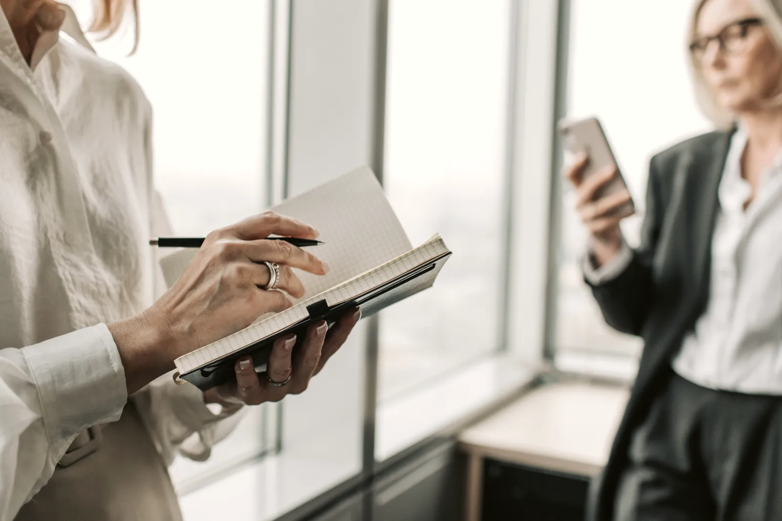 Eine Vertriebsmitarbeiterin notiert sich am hellen Bürofenster etwas, im Hintergrund schaut eine Kollegin auf ihr Telefon