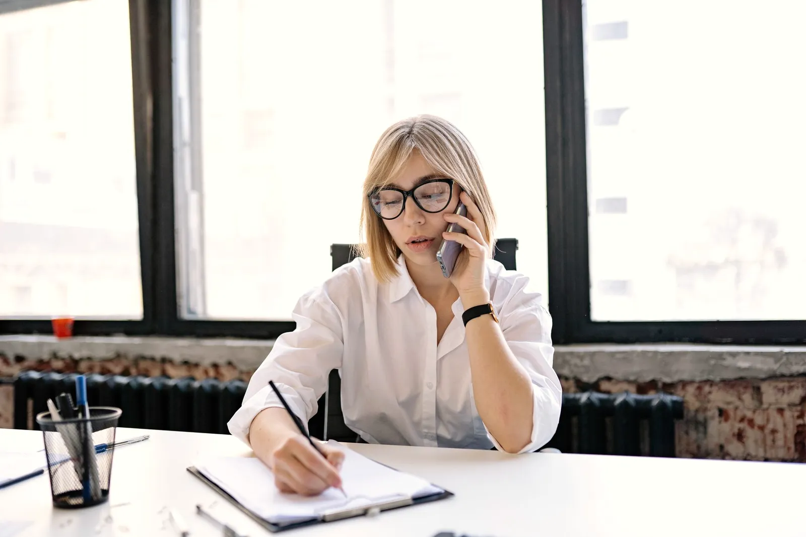 Frau am Schreibtisch in einem hellen Büro telefoniert und macht sich Notizen