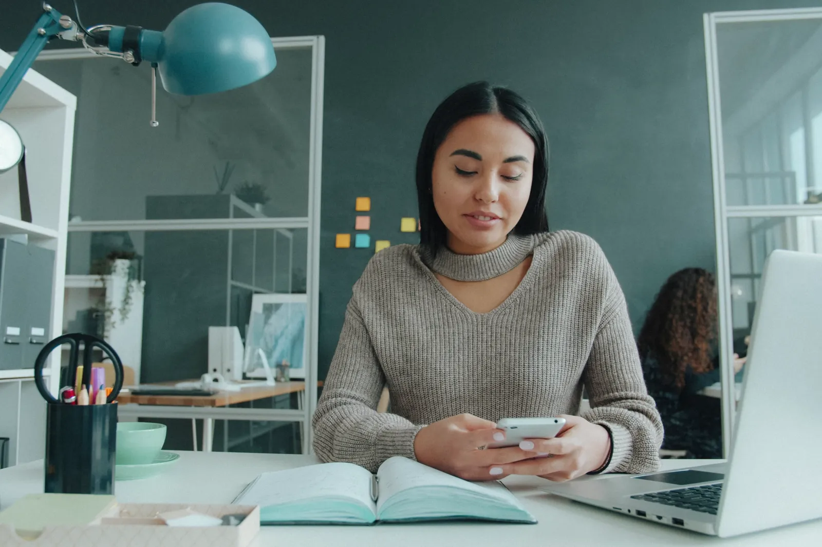 Eine Frau liest an ihrem hellen Schreibtisch eine Nachricht auf dem Smartphone, daneben Laptop und Notizbuch