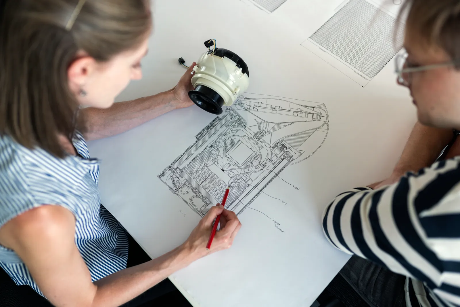 Zwei Fachleute besprechen an einem hellen Tisch eine technische Zeichnung, eine hält ein Bauteil in der Hand