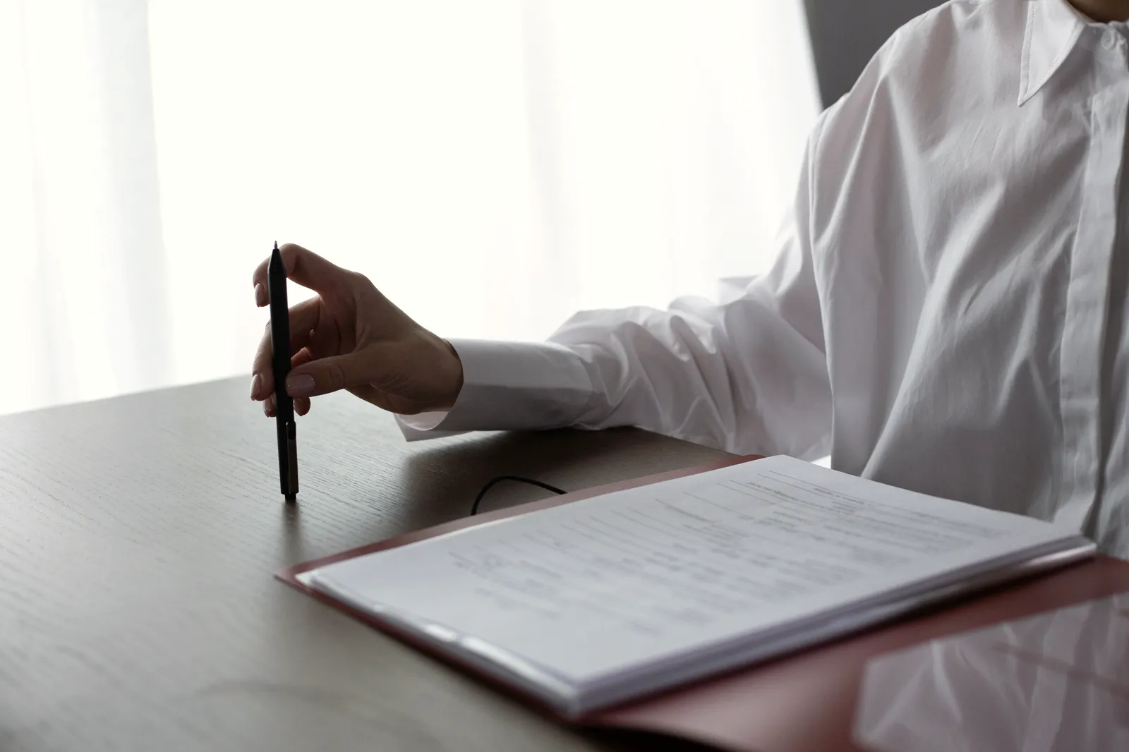 Eine Person in weißer Bluse sitzt an einem hellen Tisch mit Stift vor einem Vertrag in einer Mappe